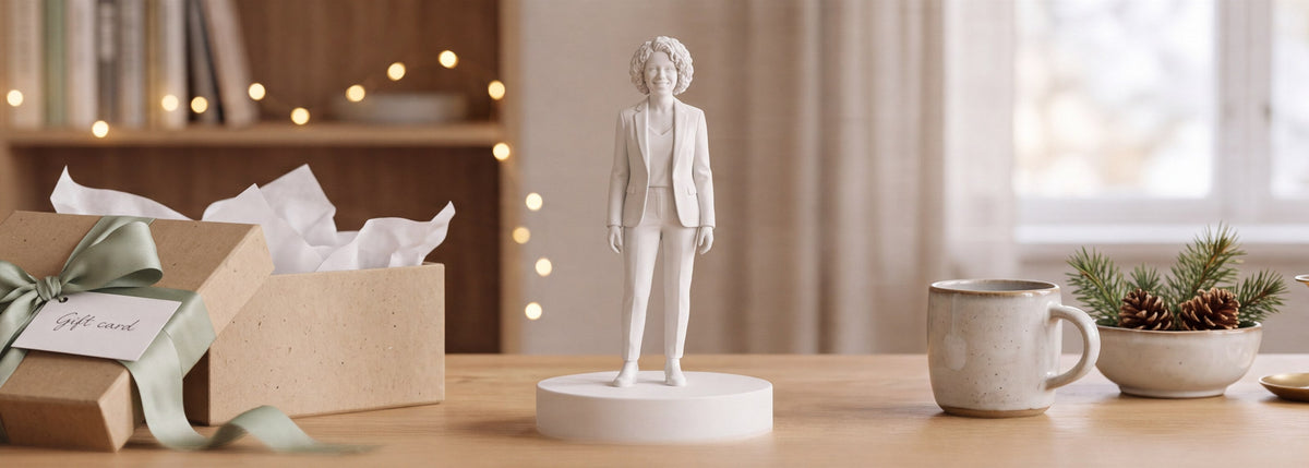 En mat hvid 3D-printet figur af en forretningskvinde på en rund sokkel står på et egetræsskrivebord mellem en åben gaveæske med salviegrønt bånd, en kaffekop, grangrene og et tændt lys, med julelys i baggrunden.
